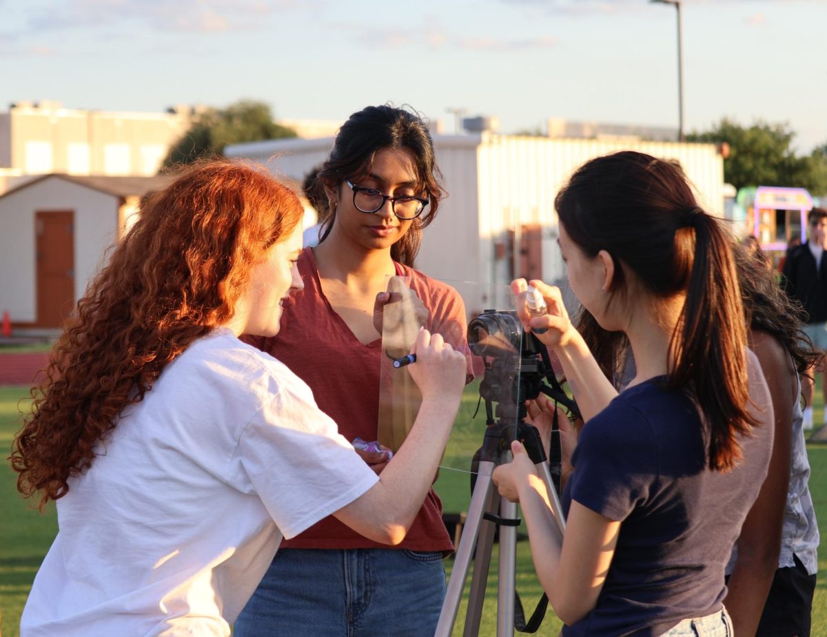 As Audrey Jesser '25 signs her name in front of the camera, Reva Chandane '26 and Karen Chen '26 hold up a clear plastic sheet. Student Council filmed multiple videos at Senior Sunset to post on their social media accounts.