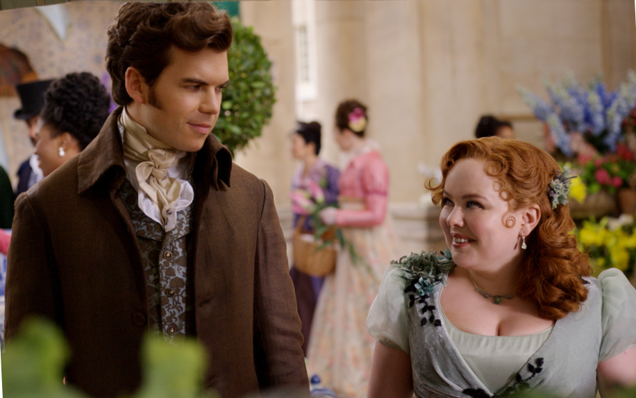 Romantic leads Penelope Featherington (Nicola Coughlan) and Colin Bridgerton (Luke Newton) steal glances at a social event. 

